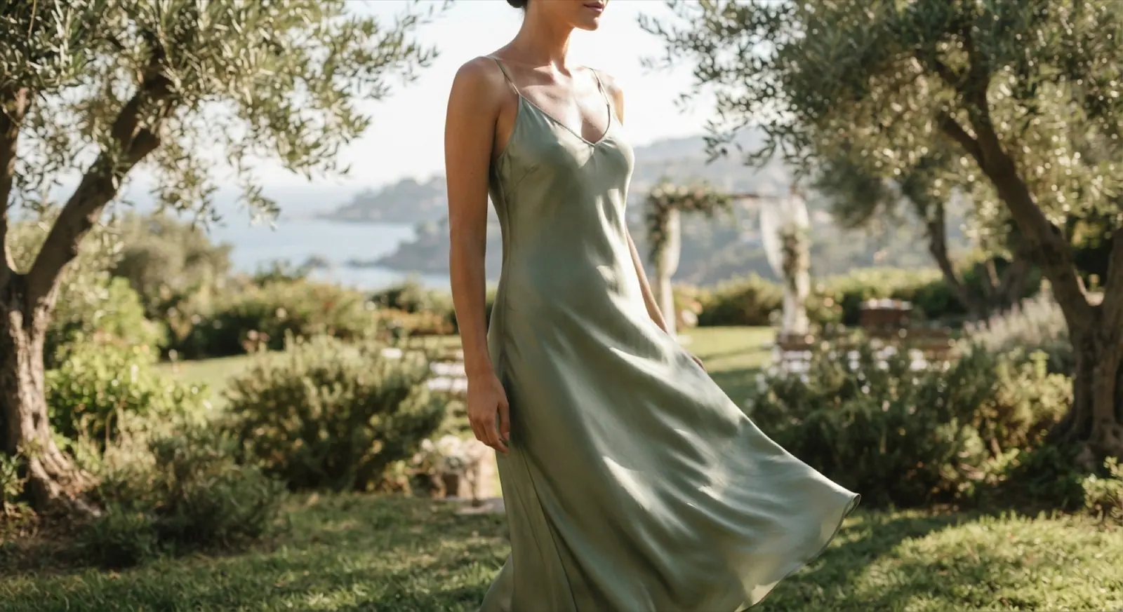 A woman in a sage slip dress walks through an olive grove above the sea