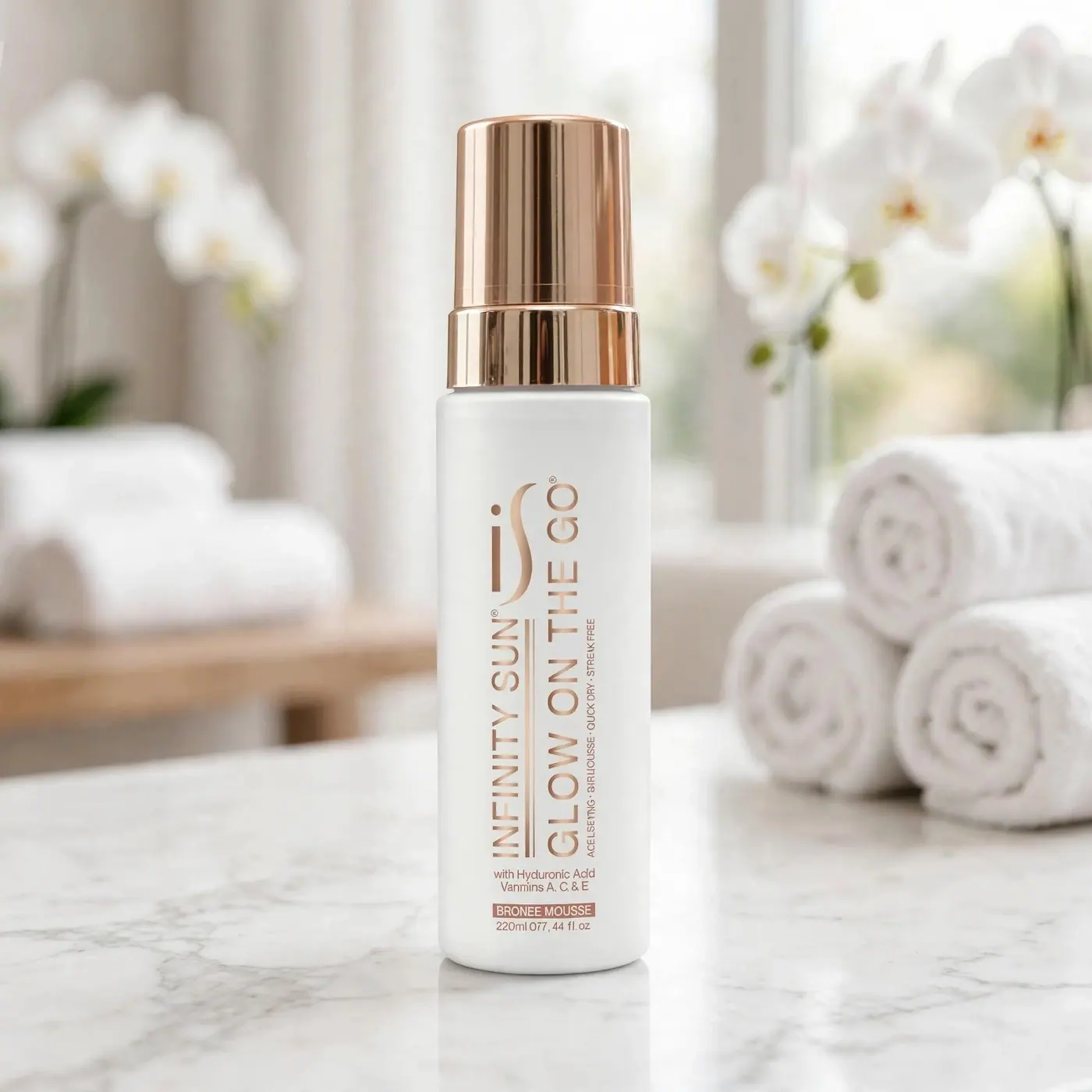 A bottle of Glow on the Go Mousse on a marble shelf beside rolled white towels