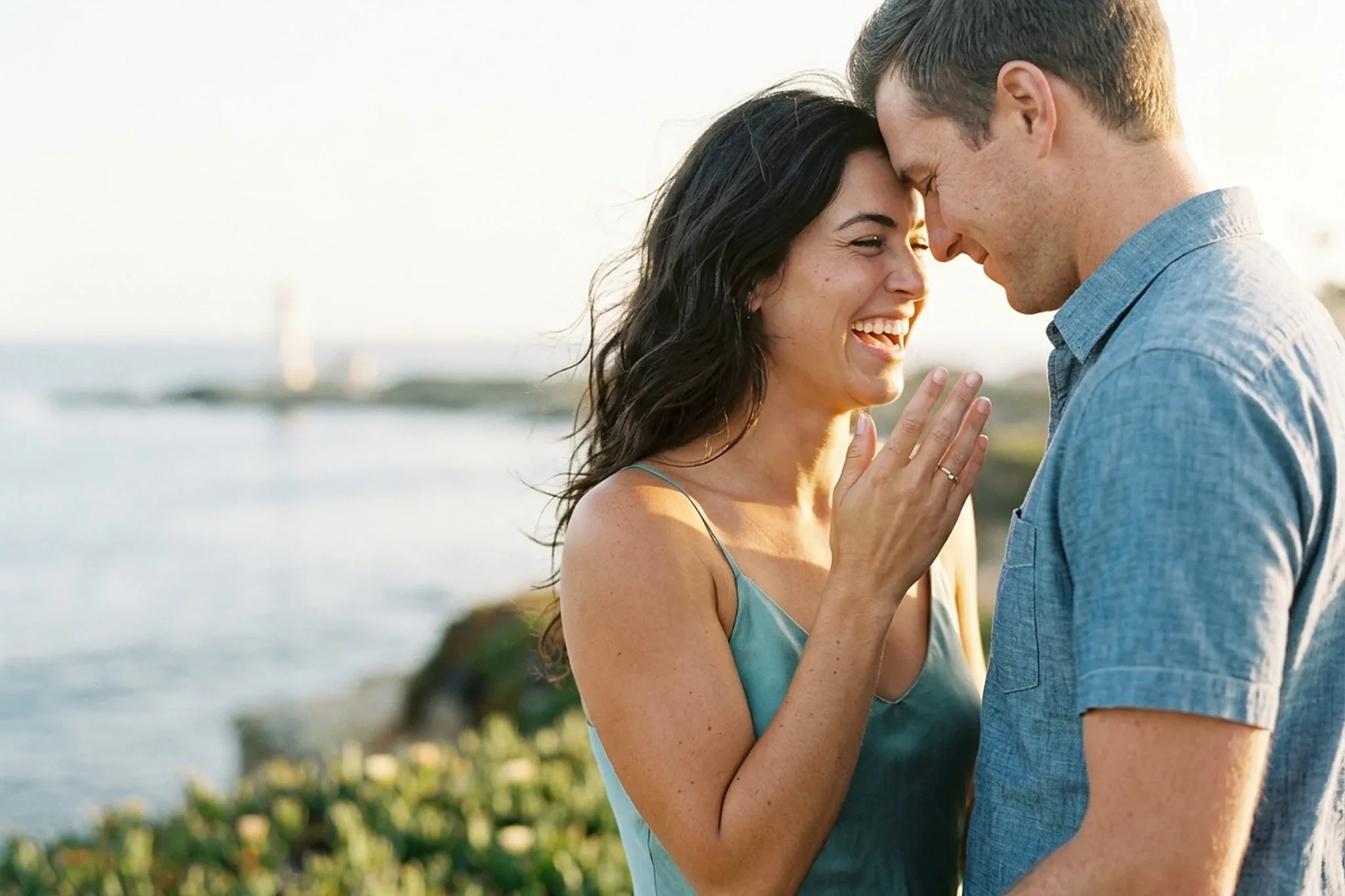 A laughing woman in a sea-glass slip dress holds up her hand to show her ring as her fiance touches his forehead to hers on a bluff above the sea