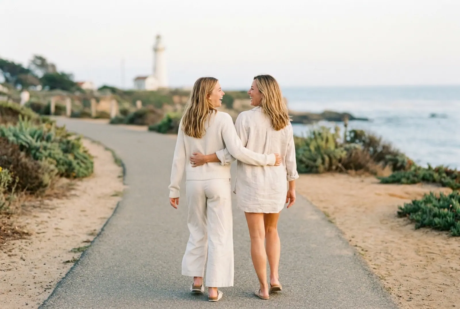 Two friends walk arm in arm along a coastal path toward a lighthouse