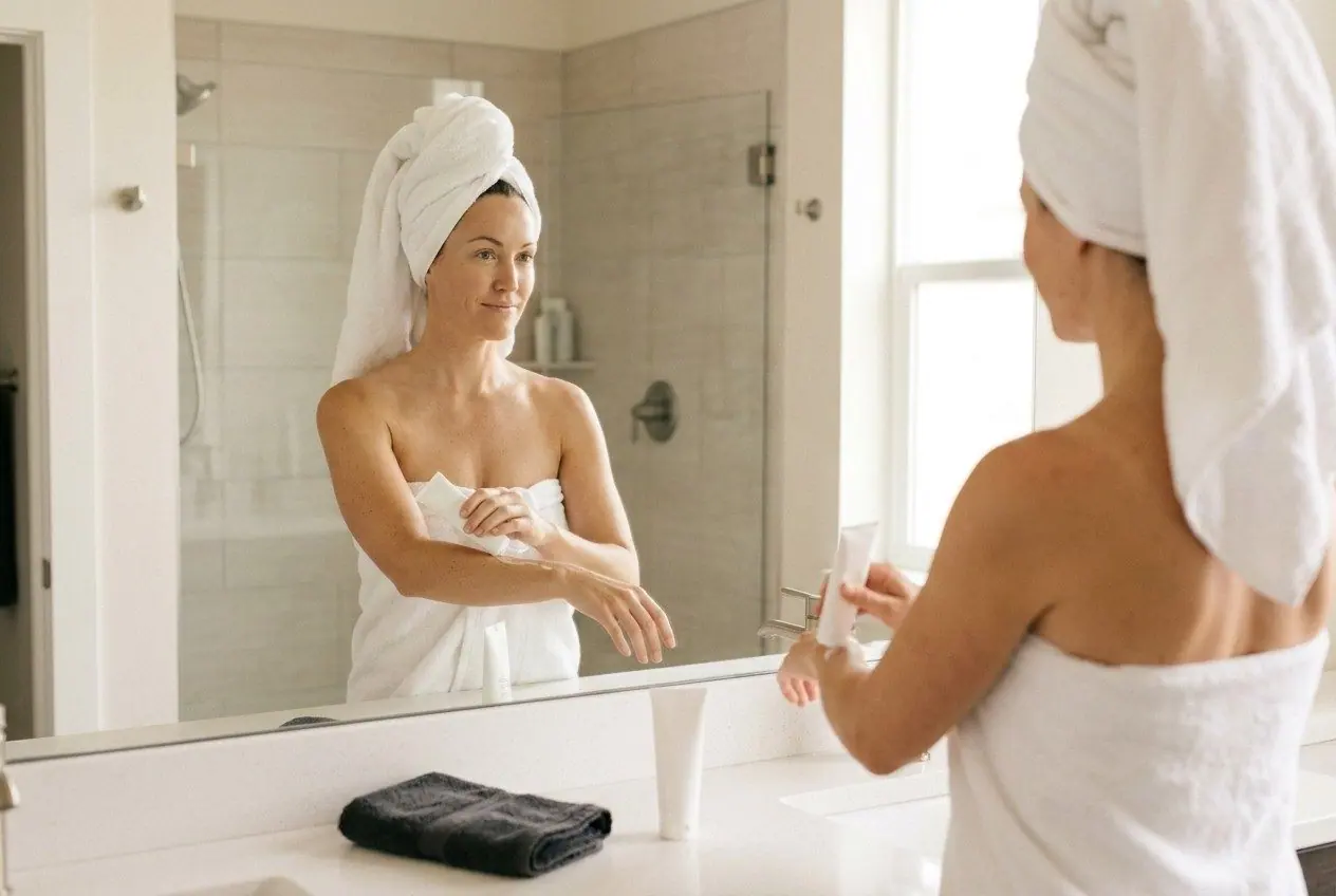 A woman wrapped in a white towel smooths cream from a tube onto her arm at the bathroom mirror