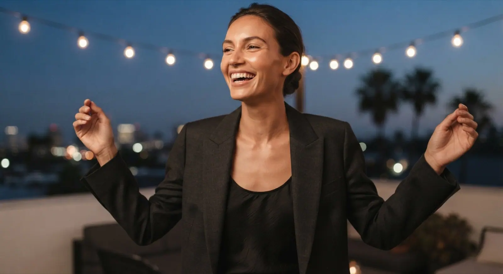 A woman in a black blazer laughs under string lights on a rooftop at night