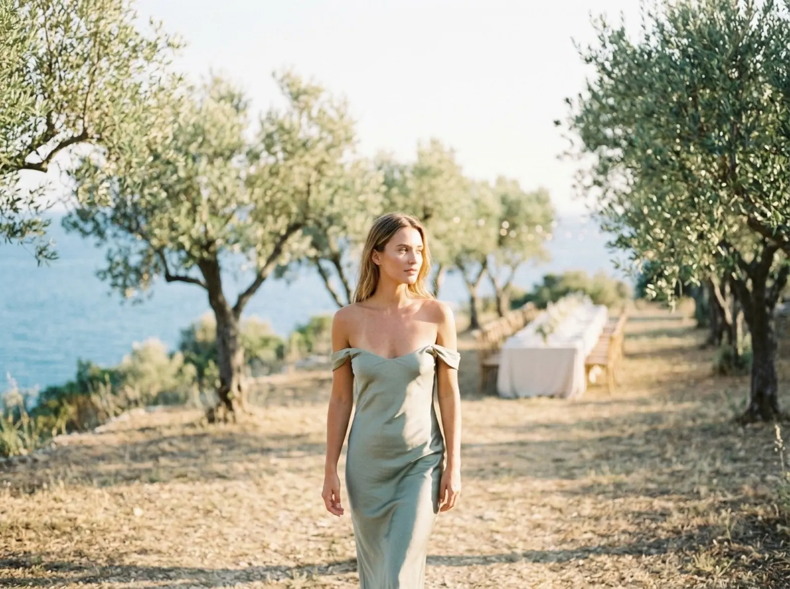 A woman in a sage slip dress walks through an olive grove above the sea