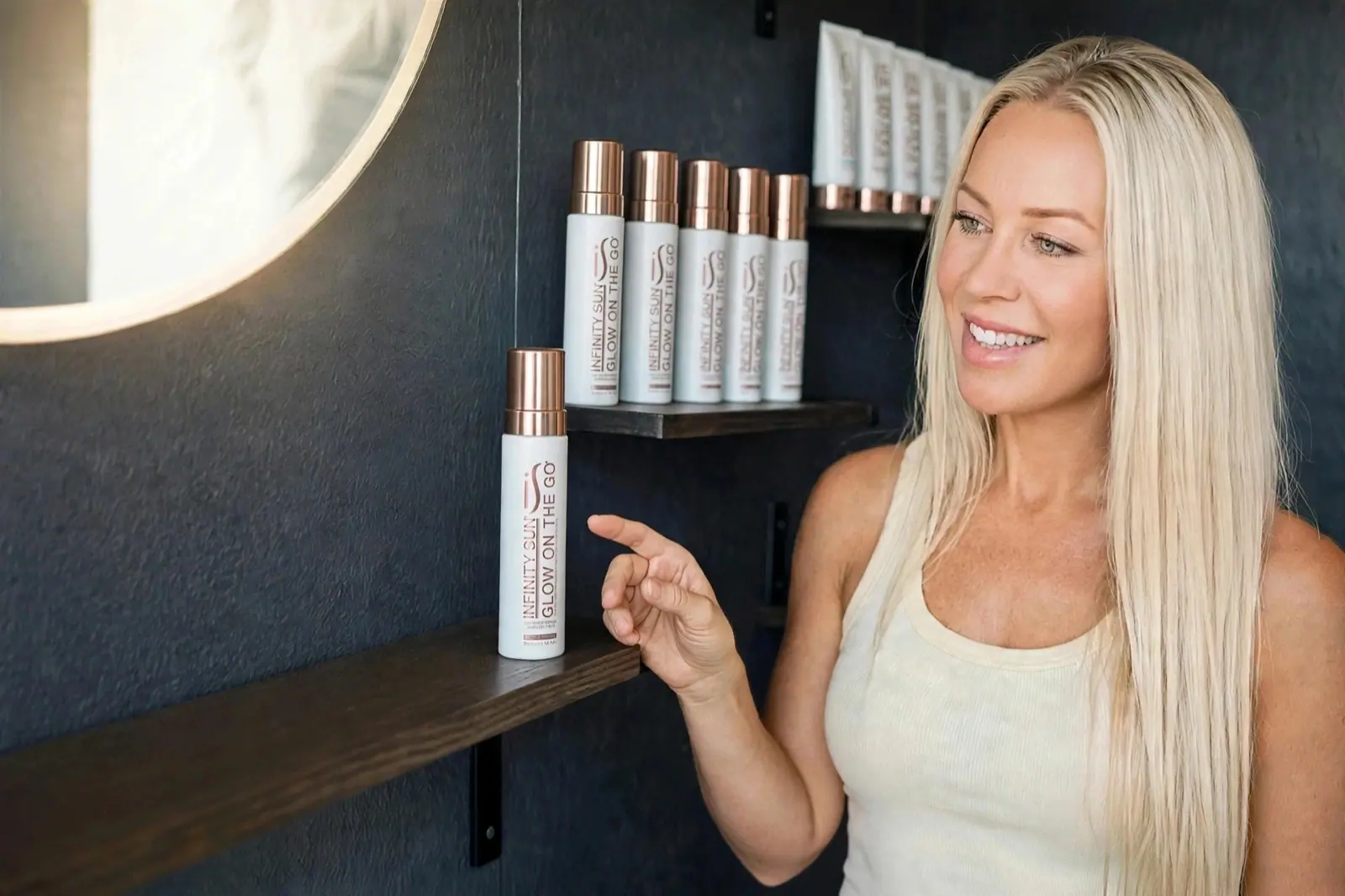 Jana at the studio shelf, pointing to a bottle of Infinity Sun Glow on the Go Mousse