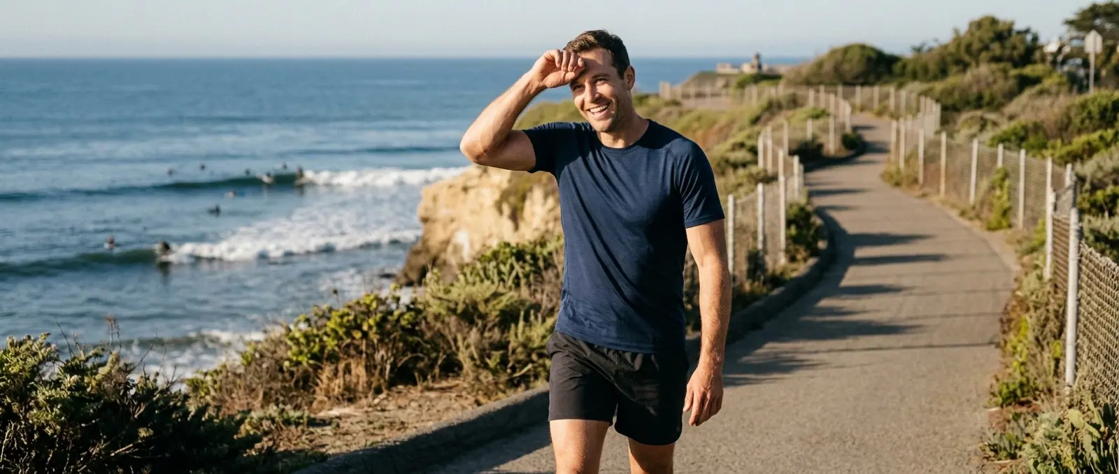 A man in a fitted navy tee walks a coastal path above the surf, shading his eyes from the sun