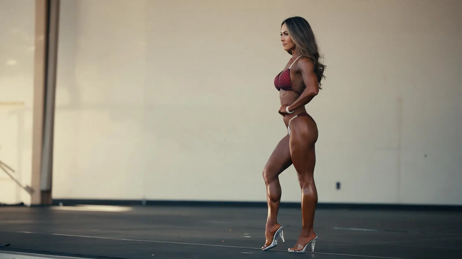 A bikini-division competitor in a burgundy competition bikini and clear heels stands in profile on a bare stage in warm light, her deep, even tan glossy across her shoulders and legs